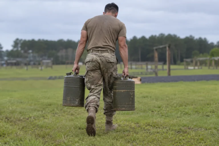 Soldier in ACUs and boots carrying two metal water cans across a field during a fitness test