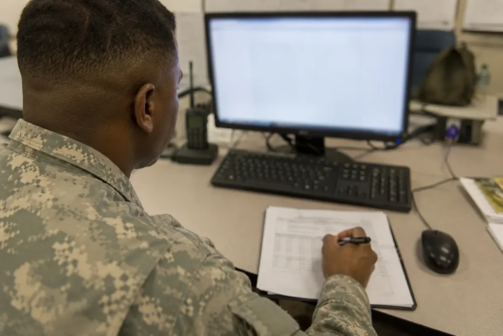 Soldier at a unit office computer entering fitness scores from a paper scorecard